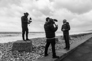 Black and white image on a beach of two men filming and another presenting
