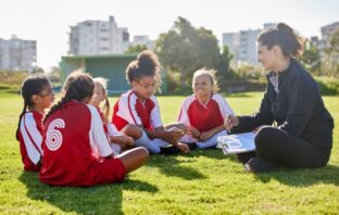 Image of young girls in a football team sitting on the field speaking to their coach