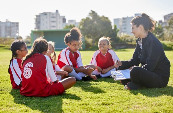 Image of young girls in a football team sitting on the field speaking to their coach