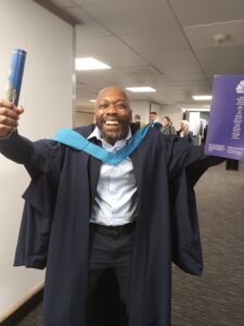 Charlie with his arms in the air and a huge smile, celebrating his degree at his graduation ceremony in OU gown.
