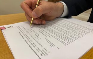 Image of a hand with a gold pen signing a document. The document being signed is the same-sex marriage regulations for Northern Ireland