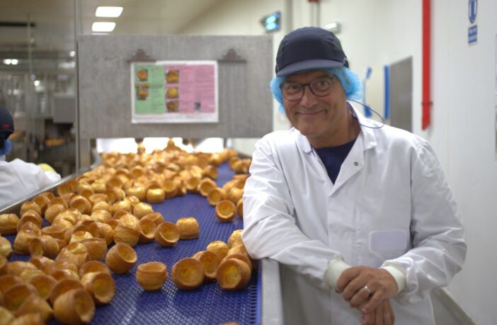 Image shows presenter Greg Wallace standing next to a production line of yorkshire puddings