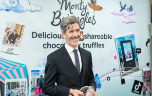 Andrew smiling, dressed in a black suit and tie, standing in front of Monty Bojangles branding, holding an award.