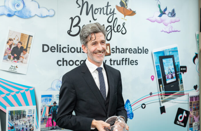 Andrew smiling, dressed in a black suit and tie, standing in front of Monty Bojangles branding, holding an award.