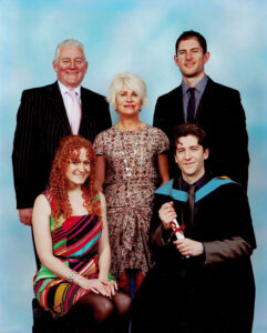 Andrew sitting on the right in his graduation gown holding a scroll with his wife Sarah sitting to the left and three family members standing behind.