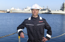 Alasdair wearing a hard hat and protective clothing in by the sea with a large ship in the background.