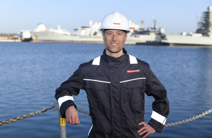 Alasdair wearing a hard hat and protective clothing in by the sea with a large ship in the background.