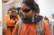 Anushree wearing an orange space suit, dark glasses and headset carrying out research. There is a colleague wearing a similar suit and glasses in the background.
