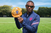 Darren Harris wearing Paralympic kit, sunglasses, facing the camera and holding a gold cap to show 100 caps for the England blind football team.