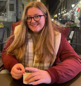 A young women with long blonde hair, wearing a red coat and scarf and eating an ice cream.