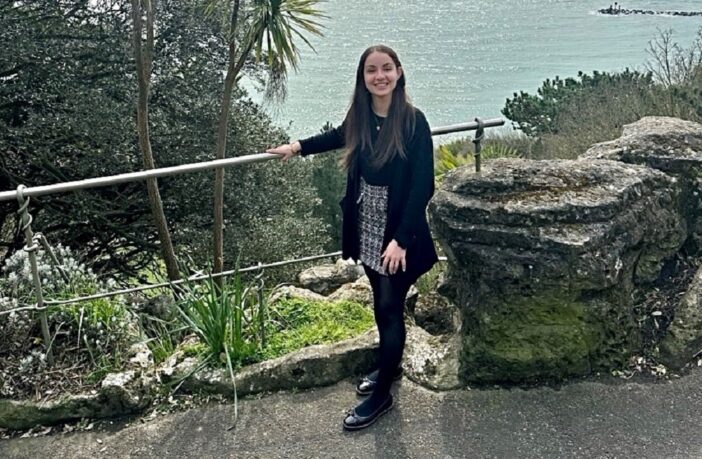 Photo of a young woman with long dark hair, standing in front of a sea view.