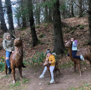 A young blonde-haired woman and her two children are sitting on model deers in a woodland setting.